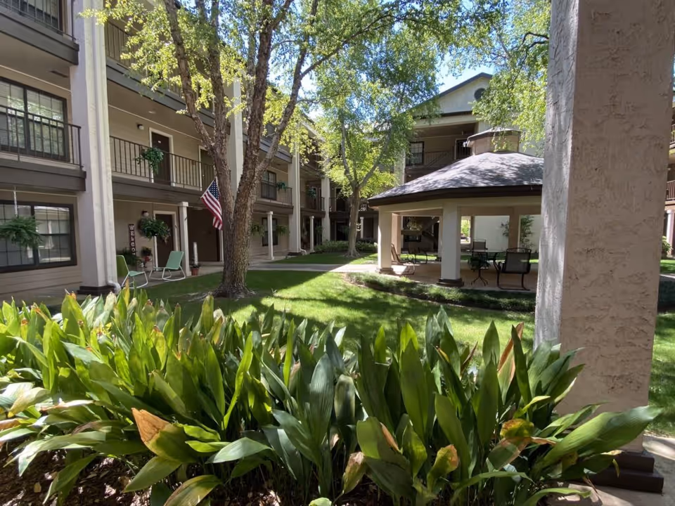 Sunlit courtyard with lush plants, a covered gazebo, surrounding multi-story residential balconies, and an American flag.