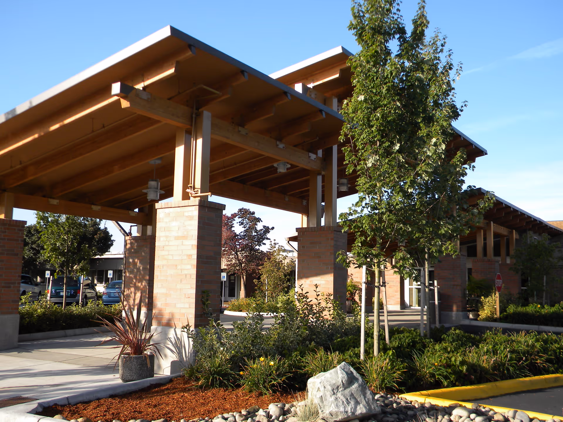 Outdoor view of a senior living facility entrance with a large wooden canopy supported by brick pillars, surrounded by landscaped greenery, trees, and a rock garden under a clear blue sky.