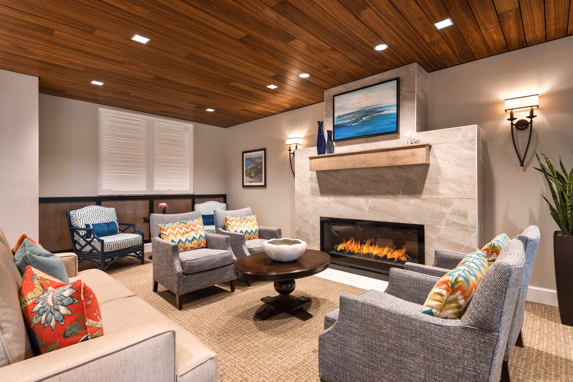 A cozy living room with a modern fireplace set in a stone wall. The room features a beige sofa with colorful patterned pillows, four gray armchairs with chevron-patterned cushions, a round wooden coffee table with a decorative bowl, and two blue patterned chairs near a window with blinds. The ceiling is wooden with recessed lighting, and there are wall sconces on either side of the fireplace. A large painting hangs above the fireplace, and a tall plant is placed in the corner.