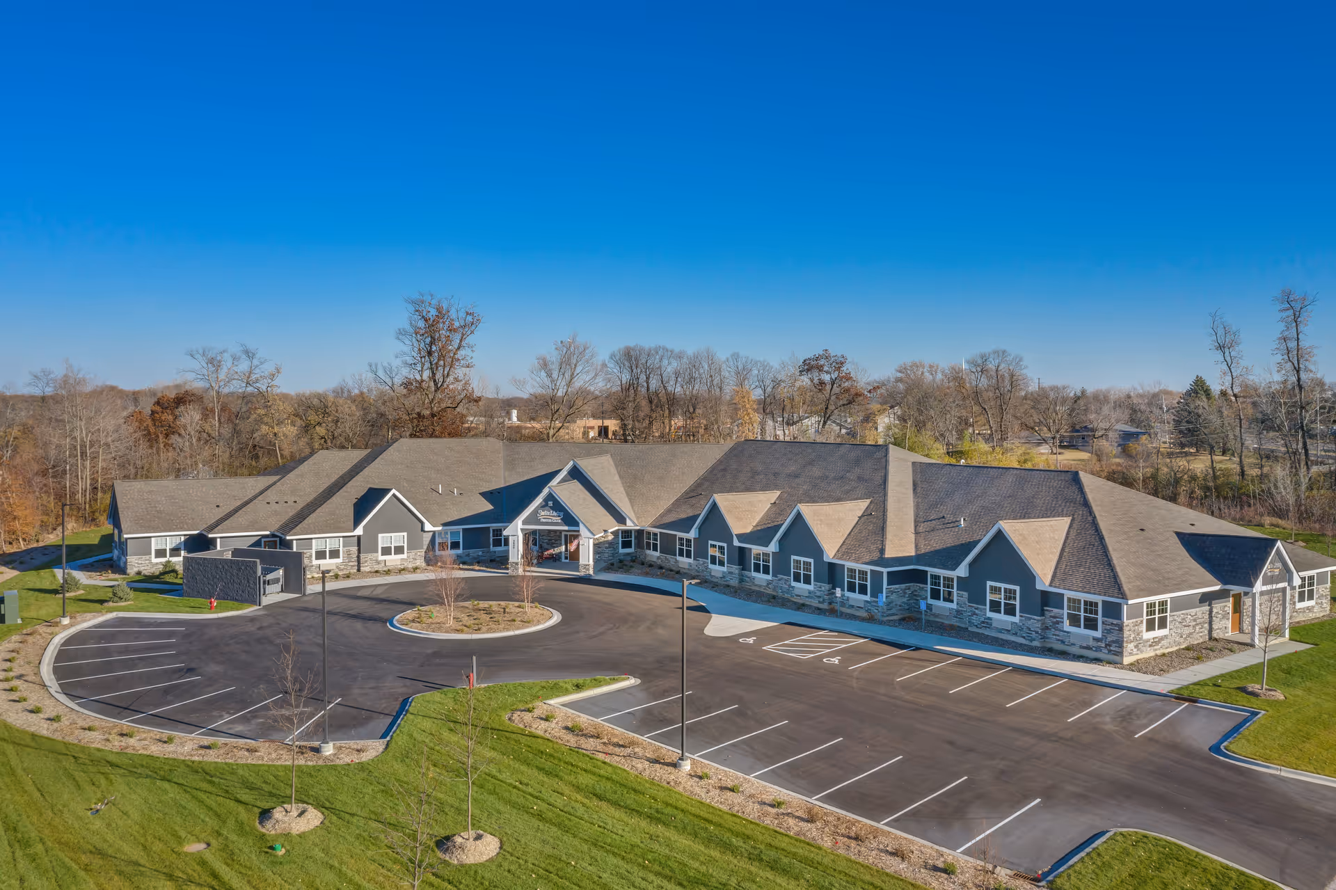 Aerial view of a single-story senior living facility building with a large parking lot and landscaped green areas surrounding it under a clear blue sky.