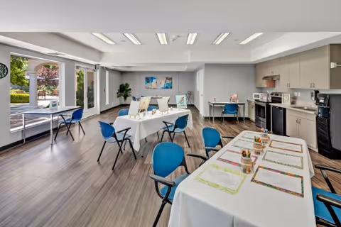 Bright multipurpose activity room with tables and easels set up for arts and crafts, blue chairs, and a kitchenette along one wall.
