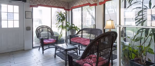 A bright sunroom with large windows and glass doors letting in natural light. The room features black wicker furniture with pink cushions, including two armchairs, a loveseat, and a glass-top coffee table. Several green potted plants are placed around the room, and a table lamp is on a side table. The floor is tiled in a light color, and the walls have a decorative border near the ceiling.