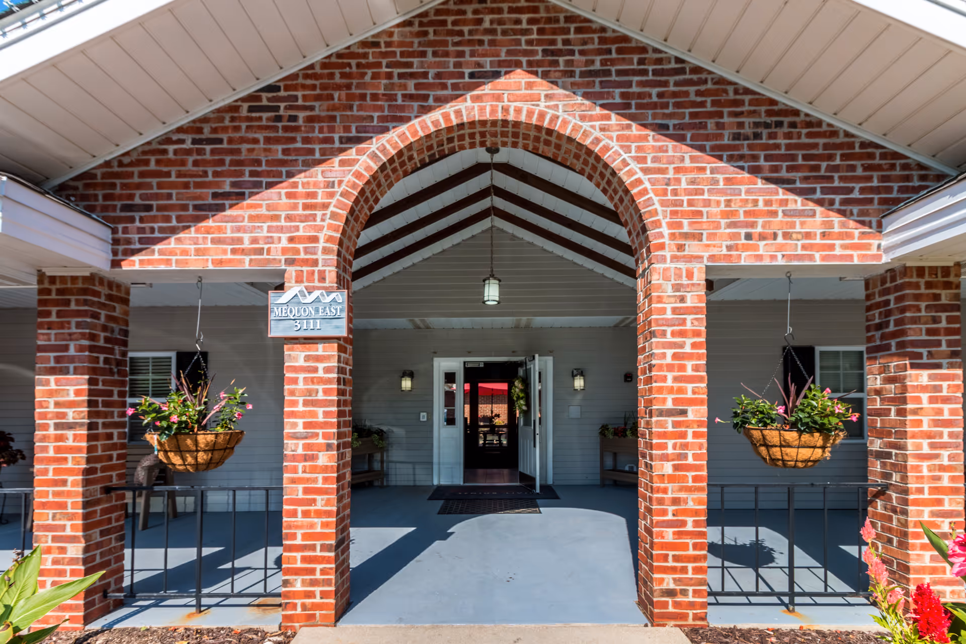 Entrance to a senior living facility with a brick archway and columns. Two hanging flower baskets with pink and green plants are on either side of the entrance. The door is open, showing a glimpse of the interior. A sign on the left brick column reads 'MEQUON EAST 3111'.