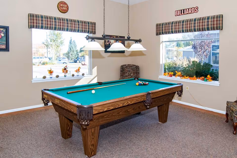 A room with a billiards table in the center, featuring a green felt surface, billiard balls arranged in a triangle, and two pool cues resting on the table. The room has beige walls, two windows with plaid valances, and decorative signs reading 'MAN CAVE' and 'BILLIARDS'. There are also floral decorations on the window sills and a patterned armchair in the corner.