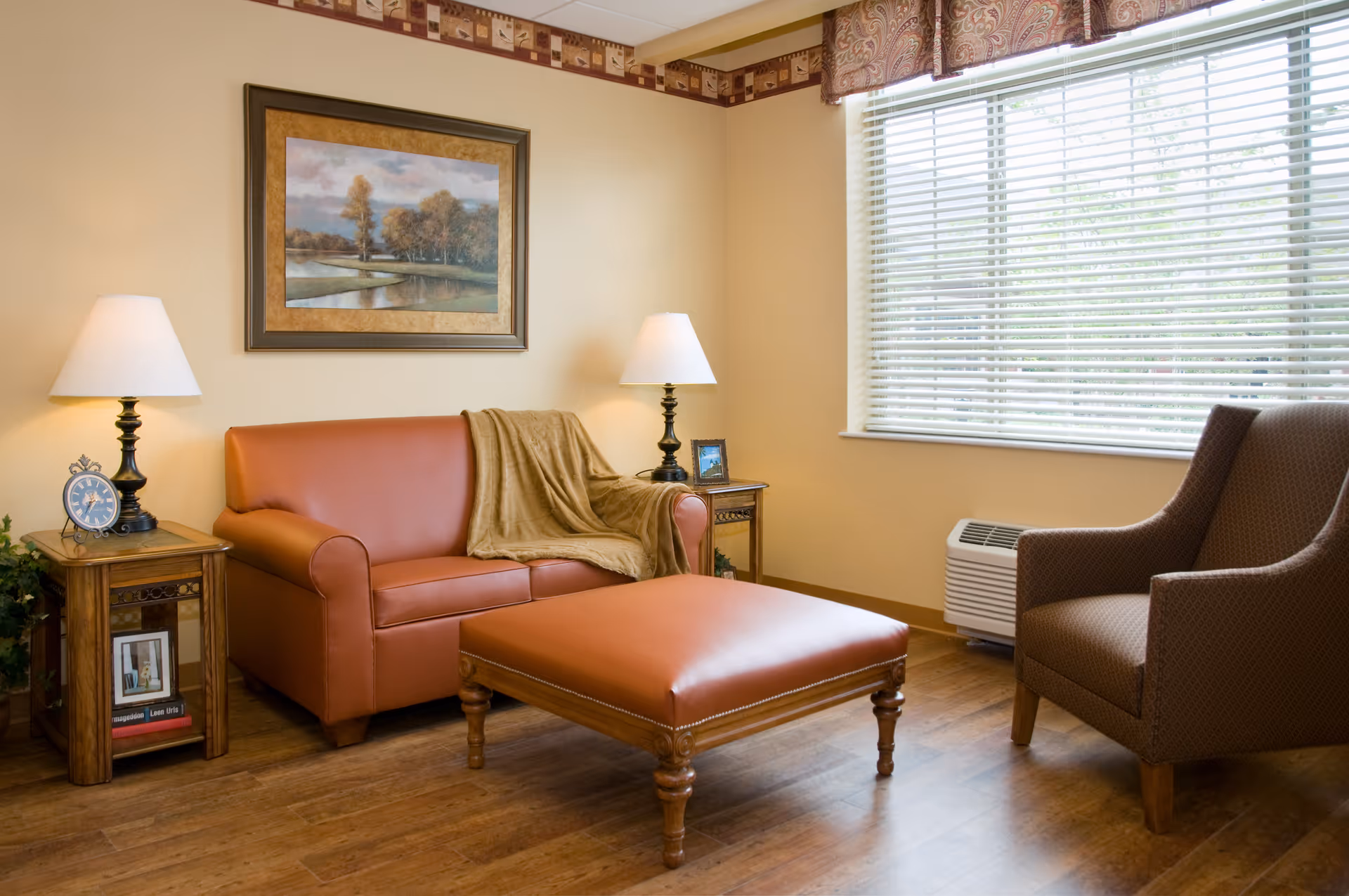 Cozy sitting room with a leather loveseat and ottoman, an upholstered armchair, side tables with lamps, framed artwork, and a large window with blinds.