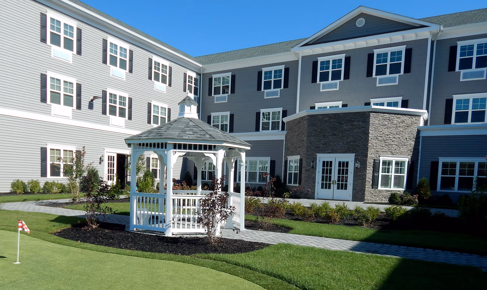 A white wooden gazebo on landscaped grounds with a small putting green in front of a three-story gray residential building.