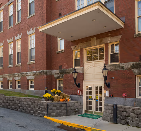 Brick senior living facility entrance with an awning, stone base, and autumn planters.