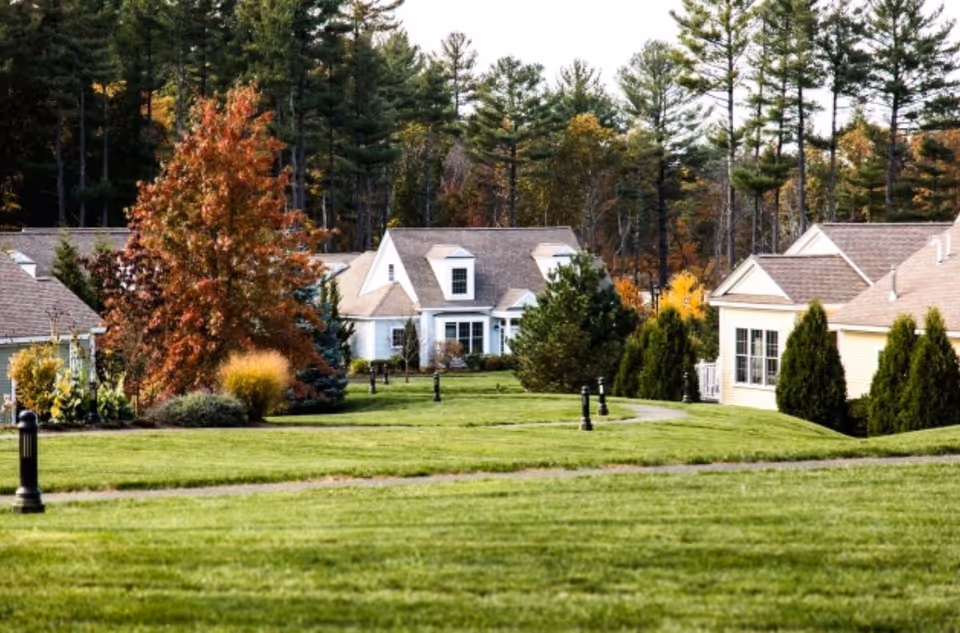 A scenic view of a senior living community with several single-story houses surrounded by green lawns, trees with autumn foliage, and a backdrop of tall pine trees.