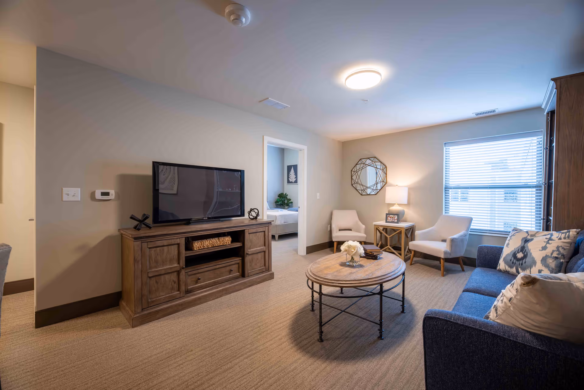 A cozy living room with a blue sofa adorned with patterned pillows, two beige armchairs, a round wooden coffee table with a flower vase, a wooden TV stand with a flat-screen TV, and a window with blinds letting in natural light. A doorway leads to a bedroom in the background.