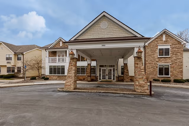 Front exterior view of a senior living facility with a covered entrance supported by stone pillars, beige siding, and multiple windows under a partly cloudy sky.