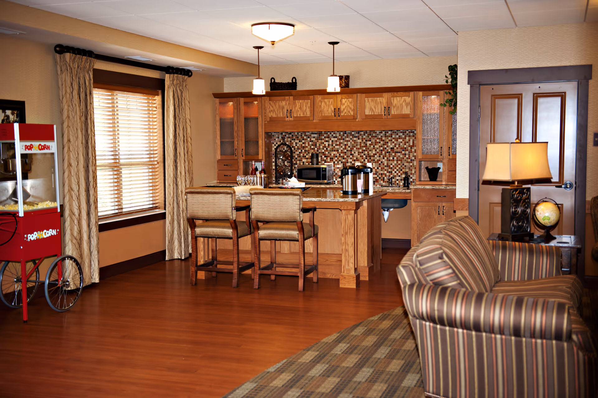A cozy interior space featuring a small kitchen area with wooden cabinets, a mosaic tile backsplash, and a kitchen island with two chairs. To the right, there is a striped sofa and a side table with a lamp and a globe. On the left side, there is a vintage-style popcorn machine near a window with beige curtains and wooden blinds. The room has warm wooden flooring and a neutral color palette.