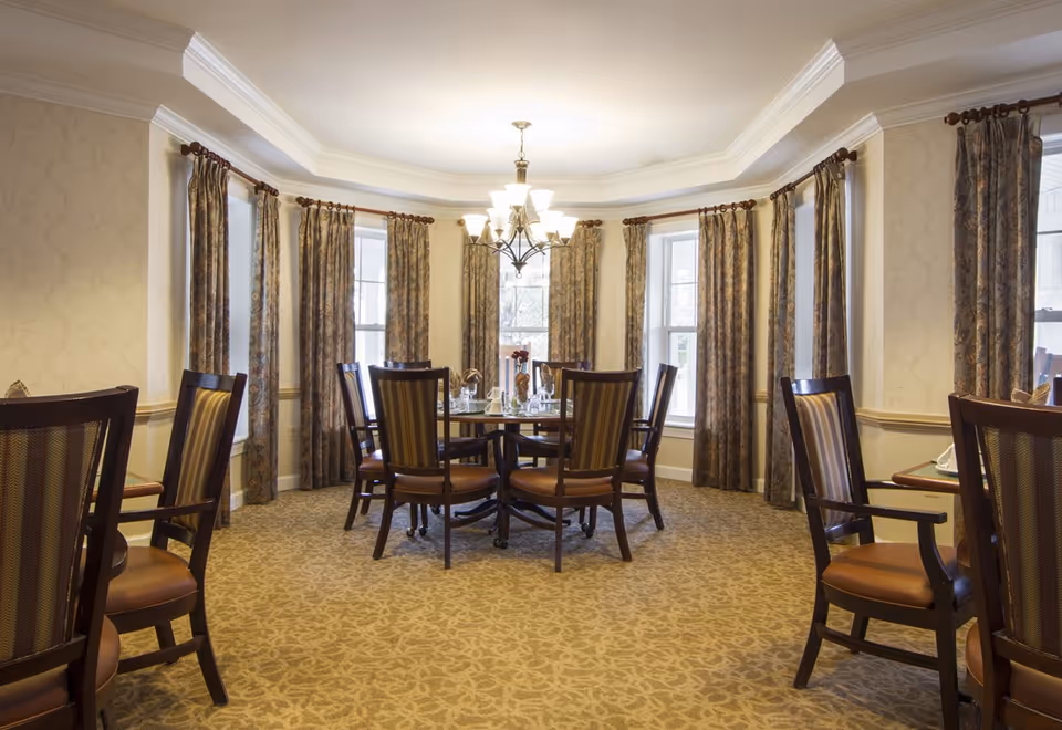 Elegant dining room with a round table and wooden chairs under a chandelier in front of bay windows with patterned curtains.