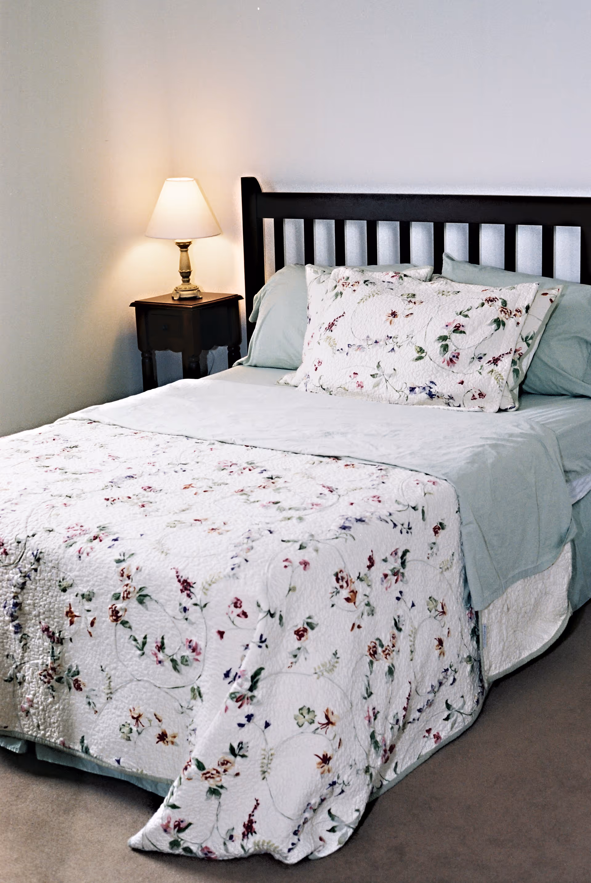 A neatly made bed with a floral quilt and pillows next to a nightstand topped with a lit table lamp.
