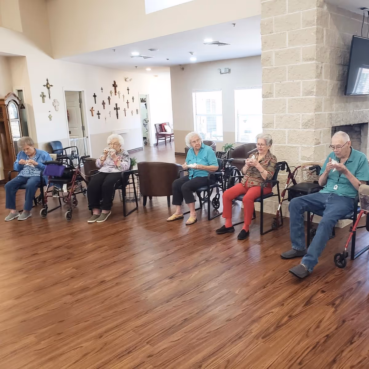 A group of five elderly individuals sitting in chairs arranged in a semi-circle in a spacious room with wooden flooring. Some have walkers nearby. The room has a stone fireplace with a mounted TV above it, and a wall decorated with multiple crosses. Large windows allow natural light to brighten the space.