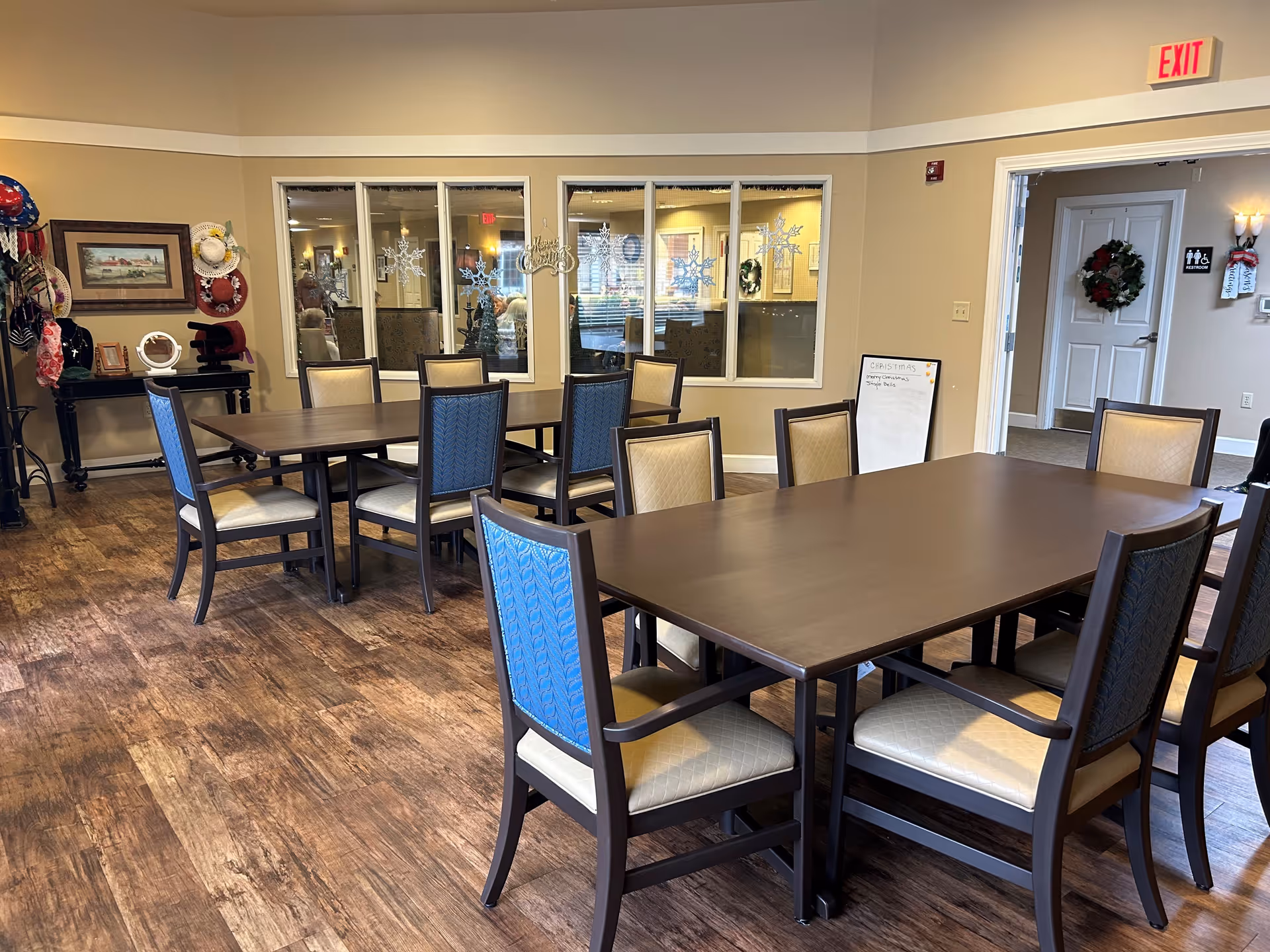 Interior view of a dining room in Cedar Ridge Alzheimer's Special Care Center with two wooden tables surrounded by chairs with beige and blue upholstery. The room has wood flooring, beige walls, and a window decorated with snowflake decals. There is a whiteboard near the doorway, a coat rack with hats and scarves, and a wreath on a door in the background.
