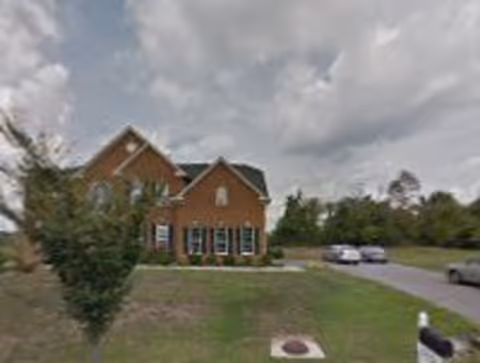 A brick residential building with multiple windows and a gabled roof, surrounded by a grassy lawn and a few trees. There is a driveway with several parked cars and a cloudy sky overhead.