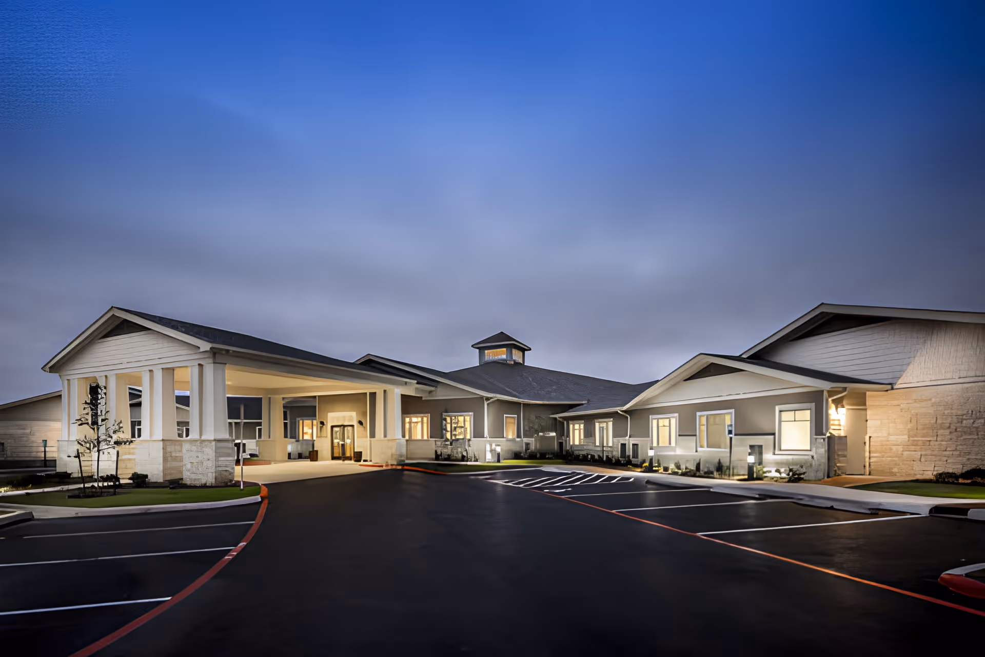 Exterior view of Chapters Living of San Antonio building at dusk with illuminated windows and a covered entrance. The parking lot in front is empty and the sky is cloudy with a blue hue.