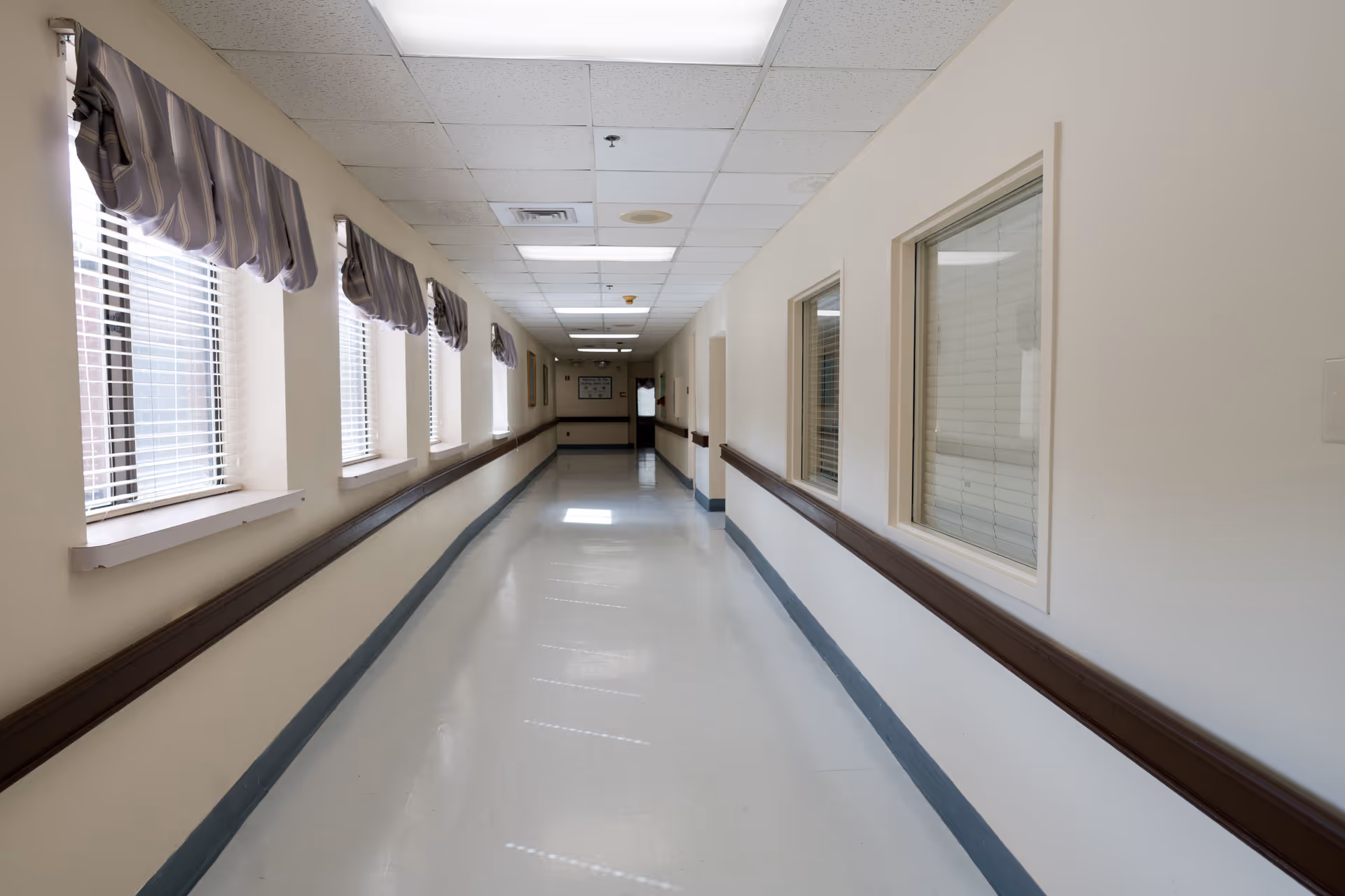 A long, clean hallway in a healthcare facility with windows on the left side covered by striped valances and interior windows on the right side. The hallway has a white floor, beige walls, and a drop ceiling with fluorescent lights.