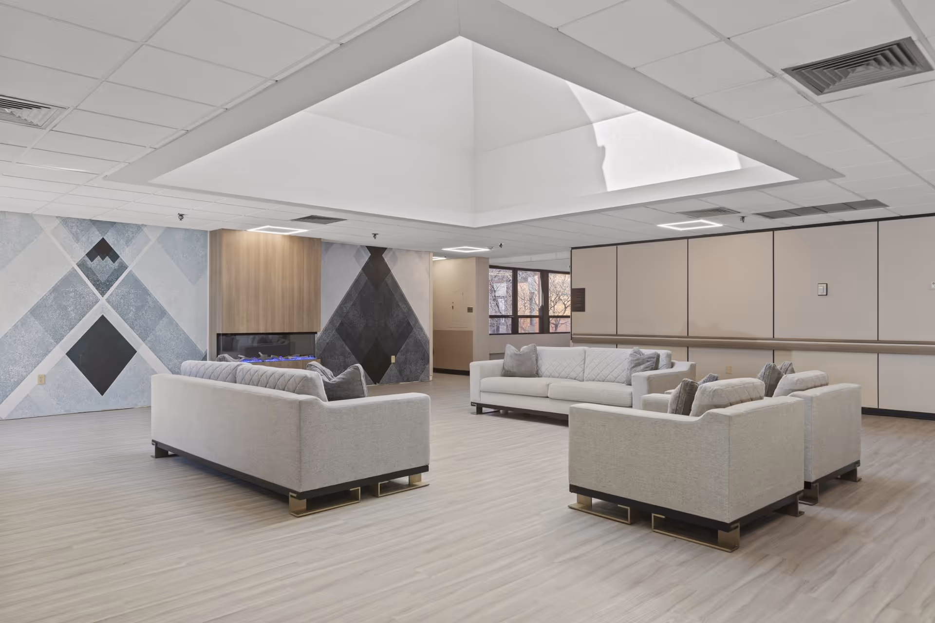 Spacious communal lounge with gray sofas arranged around a central area, geometric accent wall, fireplace, and a large recessed skylight.