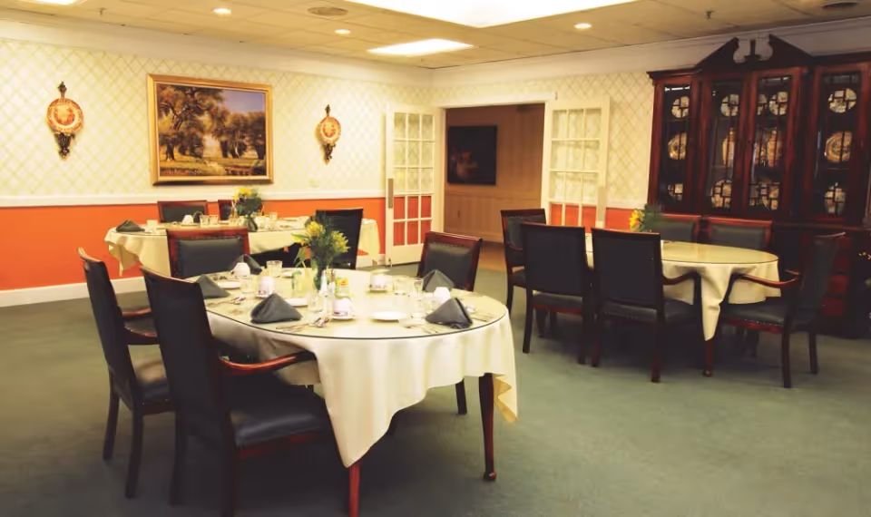 A dining room with round tables covered in white tablecloths, set with plates, glasses, napkins, and silverware. The room has several dark wooden chairs around the tables, a large wooden cabinet with decorative plates, and a painting on the wall with two ornamental wall hangings on either side.