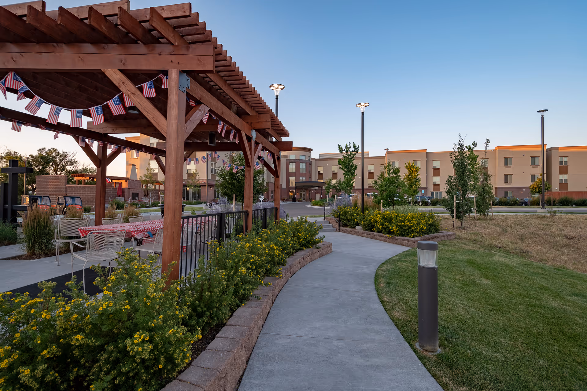 Outdoor area of a senior living facility with a wooden pergola decorated with American flag bunting, tables and chairs with red checkered tablecloths, a curved concrete walkway, landscaped bushes with yellow flowers, and a multi-story building in the background under a clear sky.