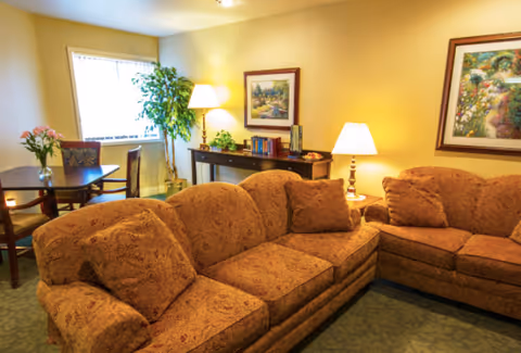 A cozy living room with two patterned brown sofas arranged in an L-shape, a wooden side table with a lamp, a console table with books and plants, framed artwork on the walls, a dining table with chairs near a window, and a potted plant in the corner.