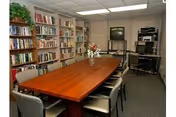 Conference-style room with a long wooden table surrounded by chairs, bookshelves along one wall, and a TV and audio equipment in the corner.