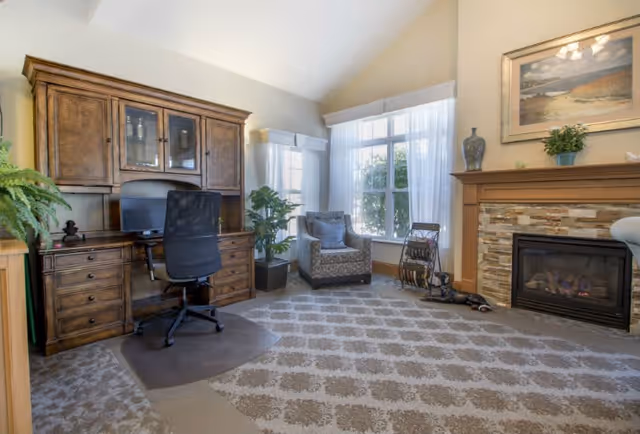 Comfortable common room with a wooden desk and hutch, an office chair, an upholstered armchair by a large window, patterned carpet, and a stone fireplace.
