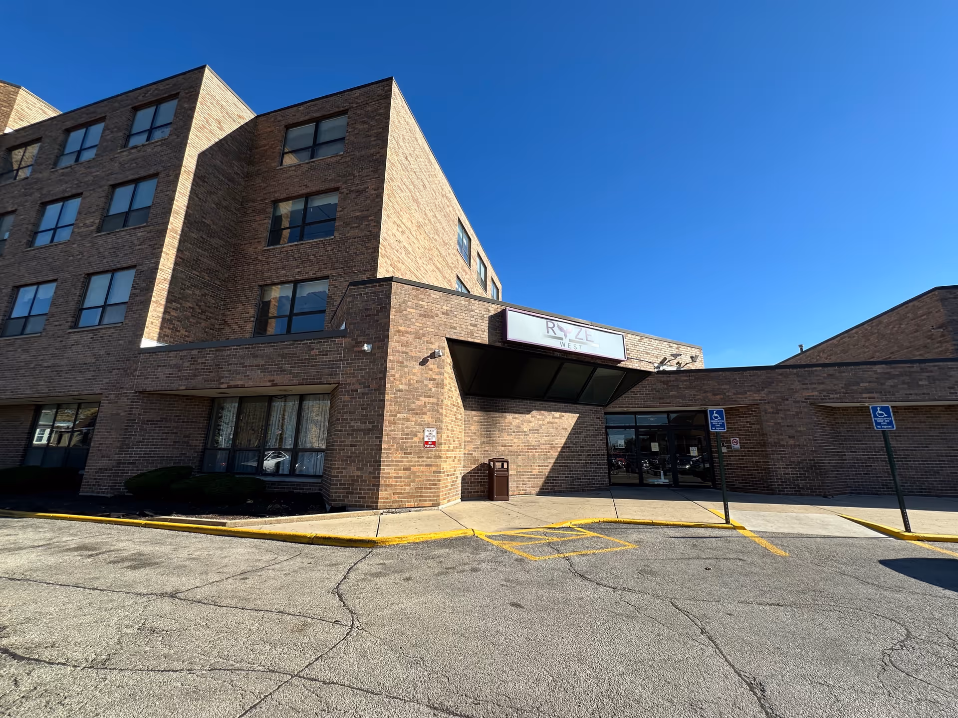 Exterior view of a multi-story brick building under a clear blue sky with a sign that reads 'RYZE West' above the entrance. The entrance has glass doors and is flanked by two handicap parking signs. The parking lot and sidewalk are visible in front of the building.