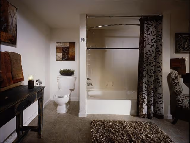 A bathroom with a white toilet, a bathtub with a floral patterned shower curtain, a dark wooden table with folded towels and a candle, a textured rug on the floor, and wall art above the toilet.