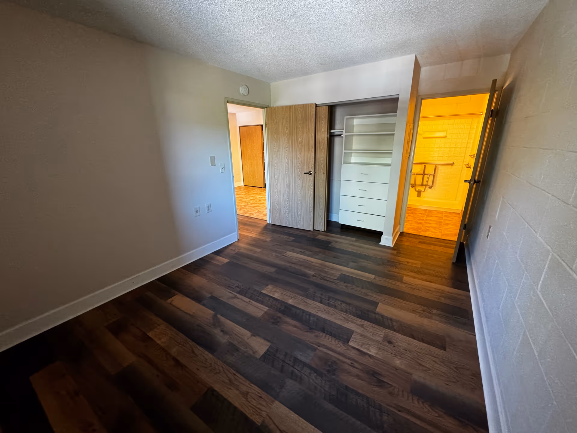 Empty room with dark wood flooring, white walls, an open closet with built-in drawers and shelves, and an open door leading to a bathroom with yellow lighting and grab bars.