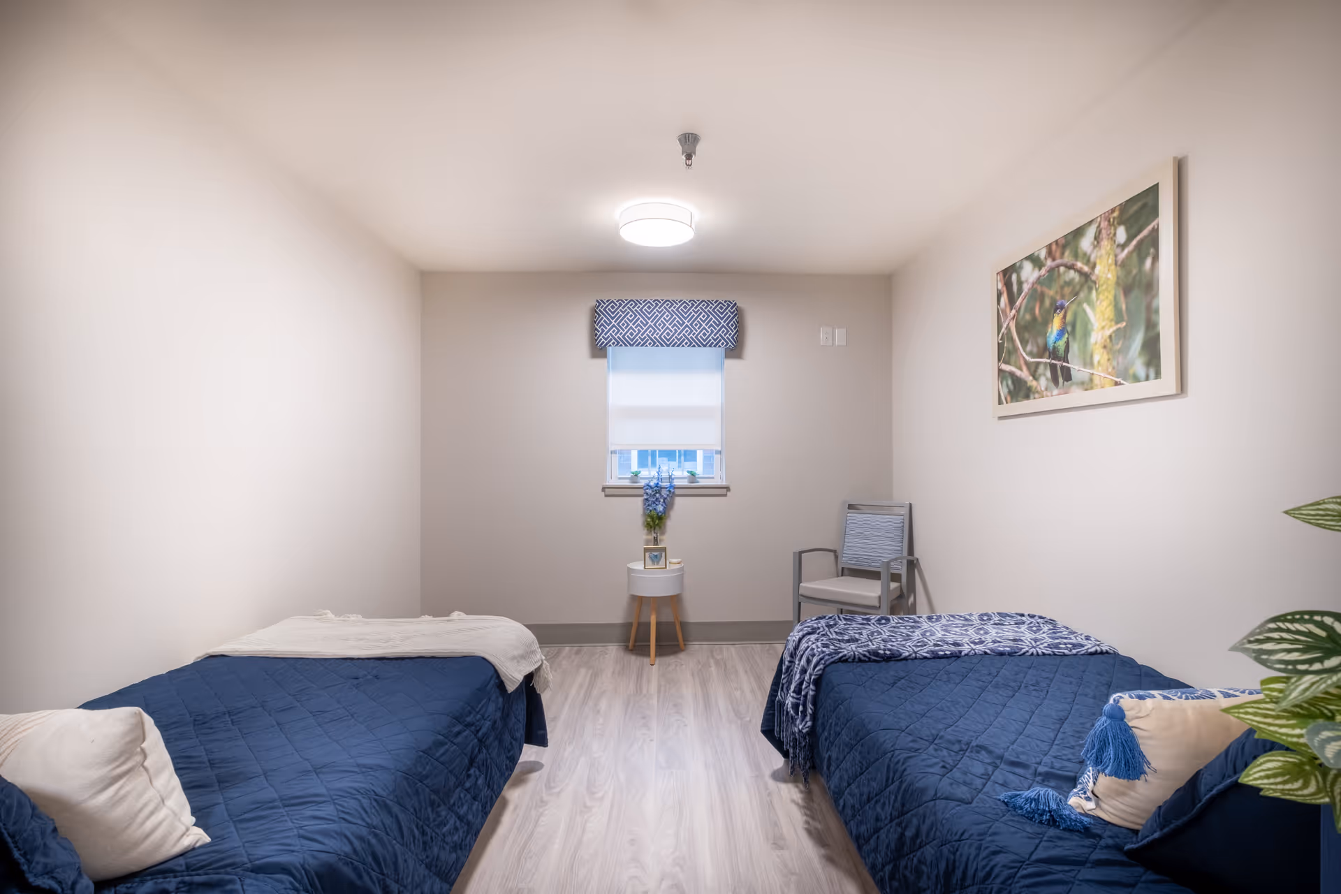 A simple, clean bedroom with two twin beds covered in dark blue quilts and white pillows. Between the beds is a small round table with a vase of blue flowers. A window with a patterned valance is centered on the far wall. A chair is placed in the corner to the right, and a framed picture of a bird on a branch hangs on the right wall. The floor is light wood, and the walls are painted a soft beige.