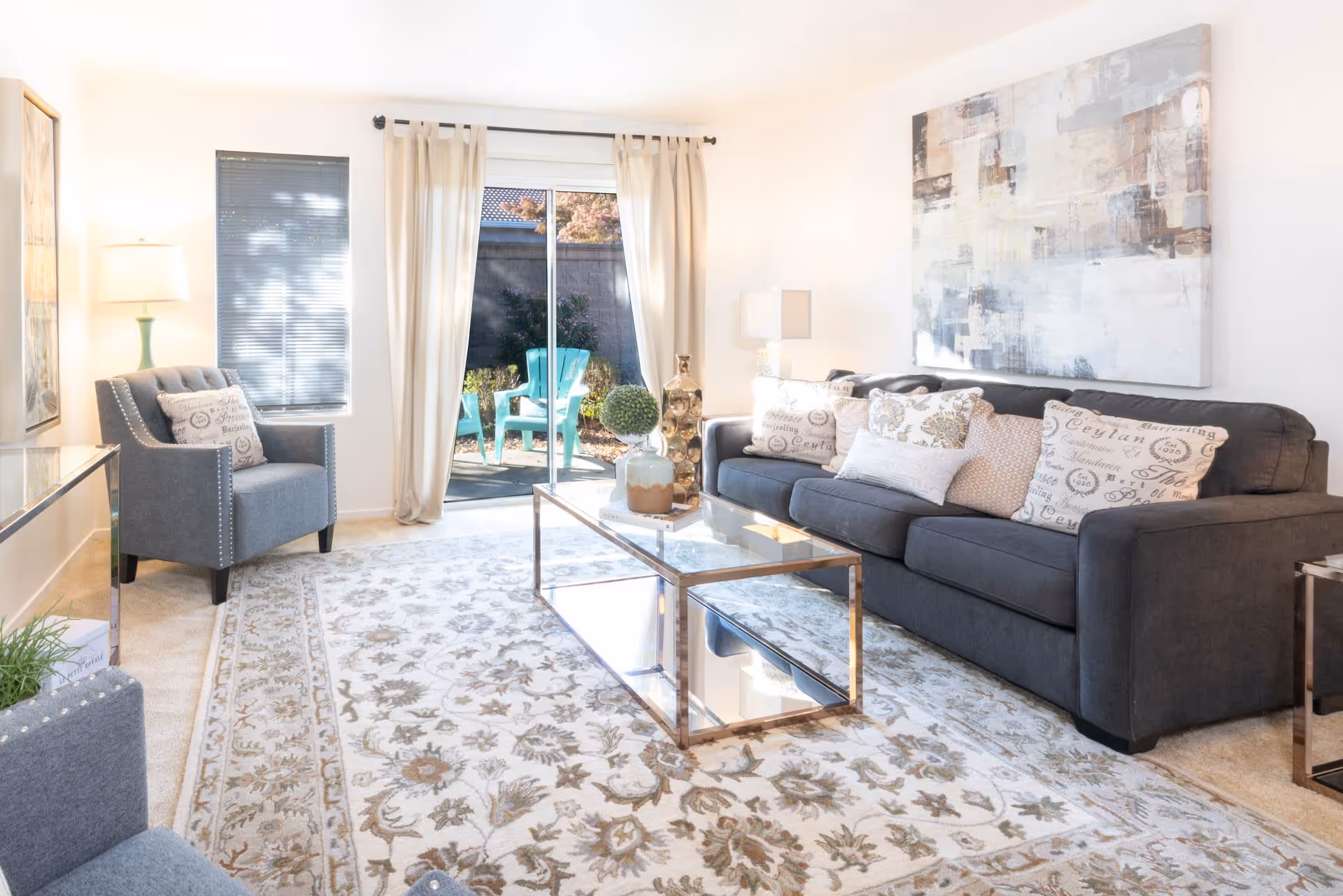 Bright living room with a dark gray sofa adorned with decorative pillows, a glass coffee table with vases and a plant, a gray armchair with a pillow, a floor lamp, and a large abstract painting on the wall. Sliding glass doors with beige curtains lead to an outdoor patio with turquoise chairs.