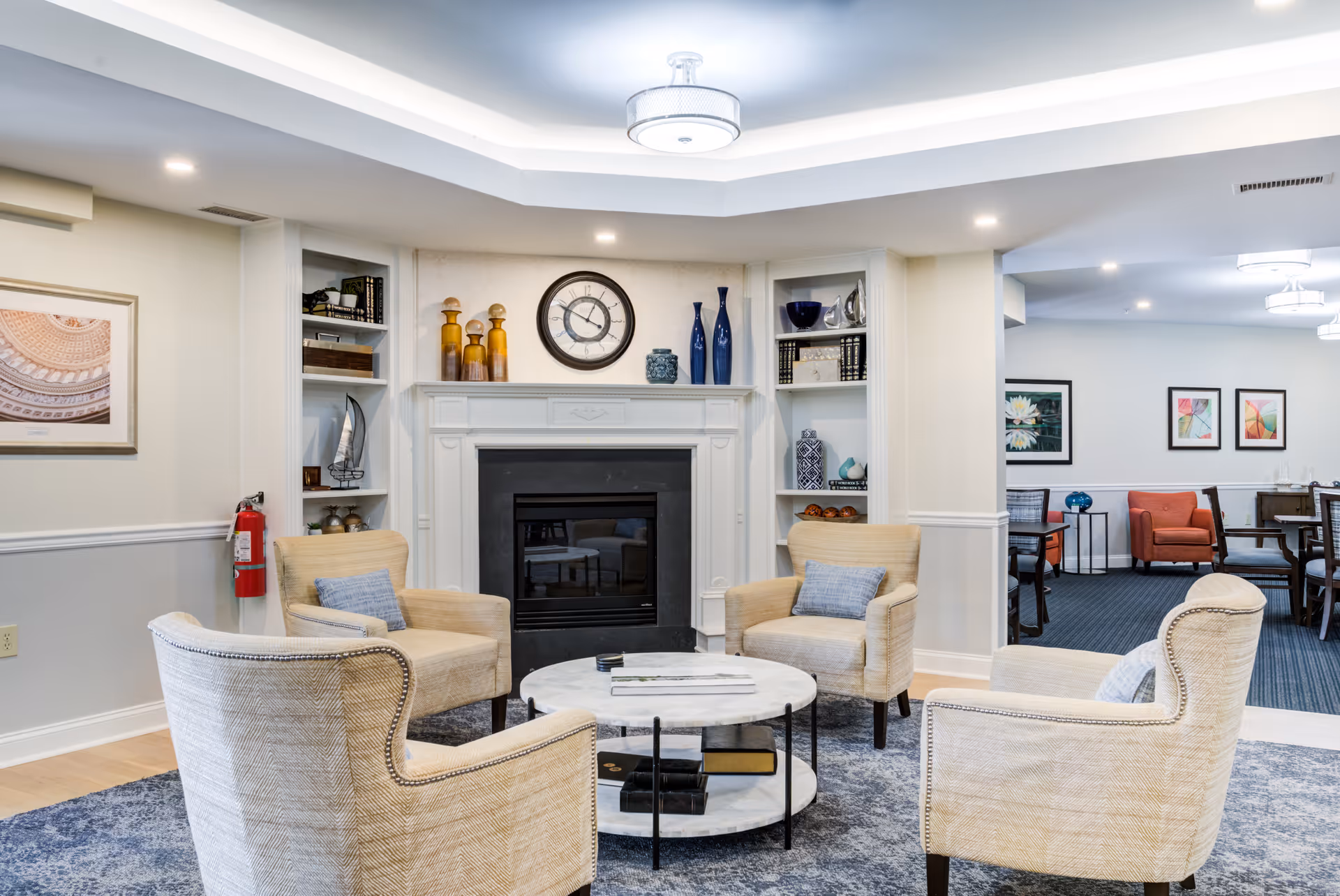A cozy senior living facility common area with four beige armchairs arranged around a round marble coffee table on a blue patterned rug. The room features a white fireplace with decorative vases and a clock above it, built-in shelves with books and decor, and soft overhead lighting. In the background, there is a dining area with tables, chairs, and framed artwork on the walls.