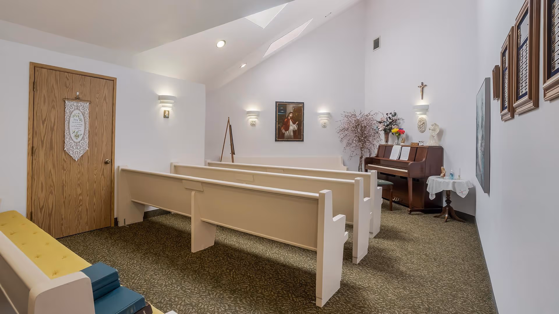 Interior view of a small chapel room with white walls and a carpeted floor. There are several white wooden pews arranged in rows facing a piano with sheet music, flowers, and religious statues on top. A religious painting hangs on the wall above the piano, and a wooden door with a decorative hanging is on the left side. The room is softly lit with wall sconces and has a peaceful atmosphere.