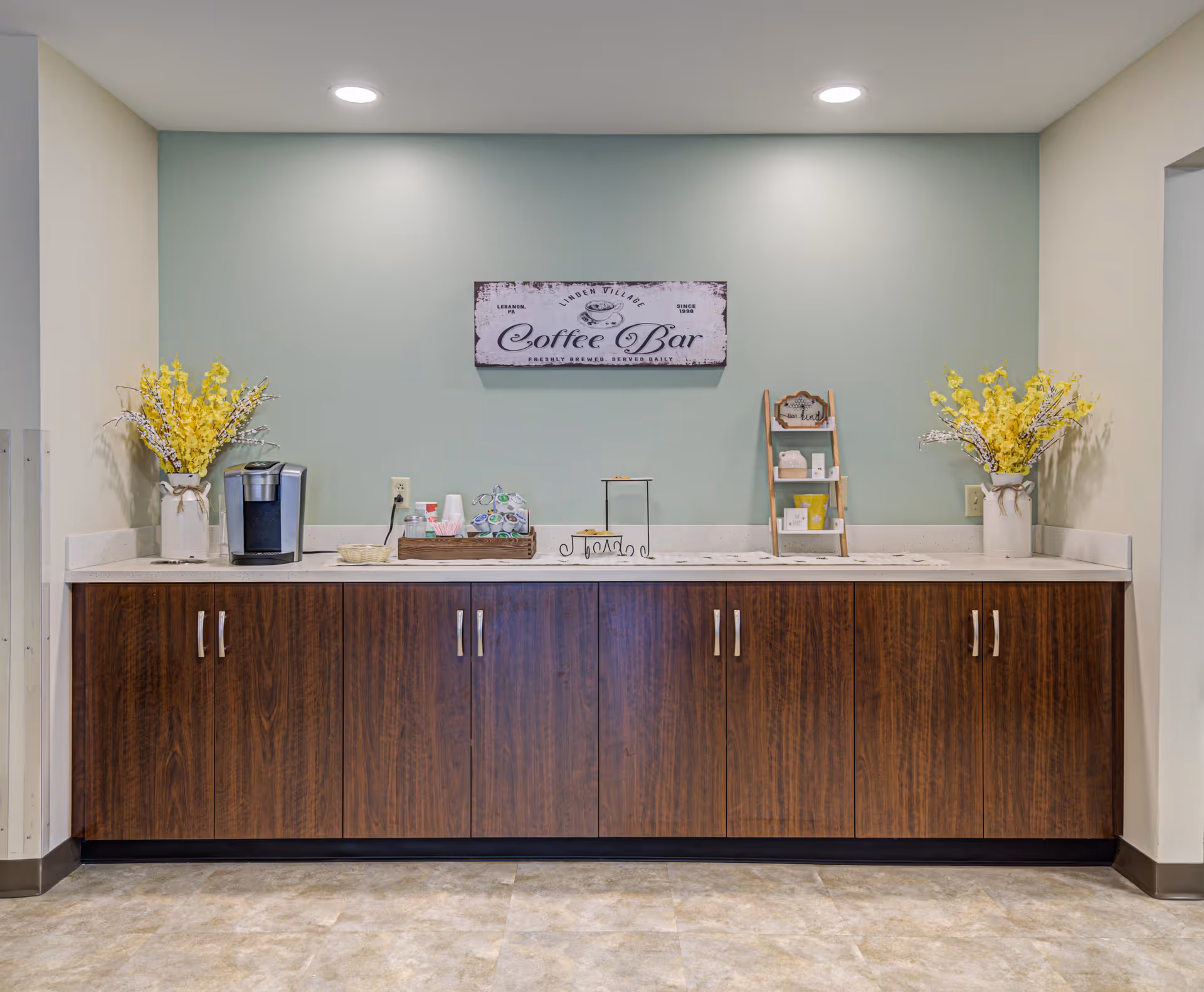 A coffee bar area with a wooden cabinet and white countertop against a light green wall. On the countertop, there is a coffee maker, a tray with coffee supplies, a small wooden shelf with various items, and two vases with yellow flowers on each end. Above the counter is a sign that reads 'Coffee Bar'.
