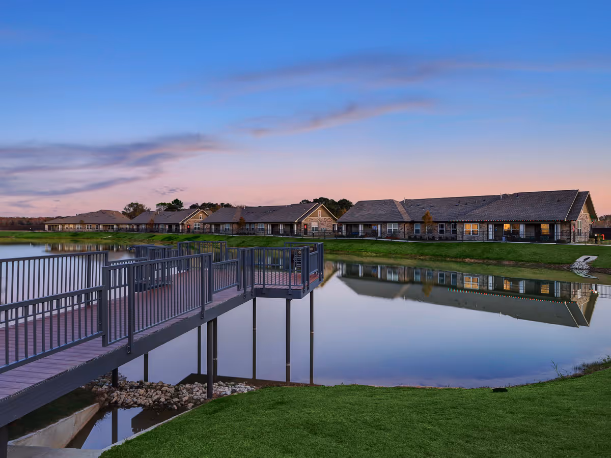 A lakeside view at dusk with a walkway dock and a row of single-story residential buildings reflected in the water.