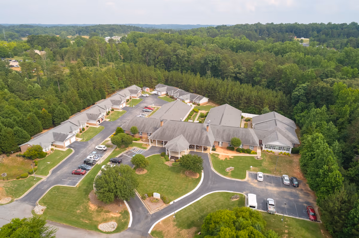Aerial view of Brookside Commerce senior living facility surrounded by dense green trees. The facility consists of multiple single-story buildings with gray roofs arranged around parking lots and driveways. Several cars are parked in the lots, and well-maintained lawns and landscaping are visible throughout the property.