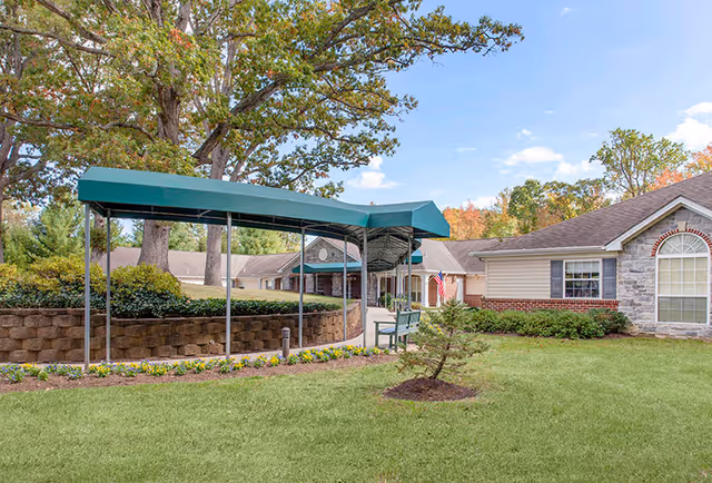 Outdoor view of Charter Senior Living of Annapolis facility showing a covered walkway with a green canopy, a bench, well-maintained lawn, small tree, flower bed, and part of the building with stone and brick exterior under a blue sky with some clouds.