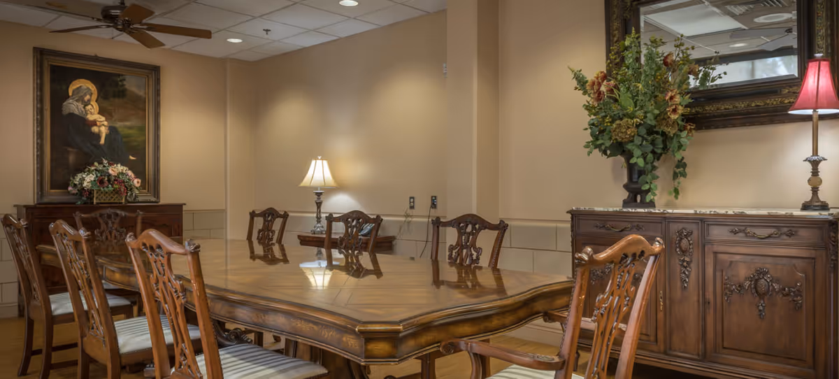 A traditional dining room with a large wooden dining table surrounded by eight intricately carved wooden chairs with striped cushions. The room features warm beige walls, a ceiling fan, a table lamp on a small side table, a large framed painting of a woman holding a child, and a wooden sideboard with a floral arrangement and a lamp with a red shade.
