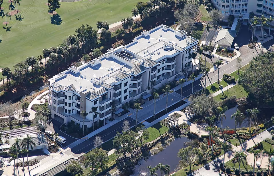Aerial view of a multi-story residential building surrounded by landscaped gardens, walking paths, palm trees, and a small pond, with a golf course visible in the background.