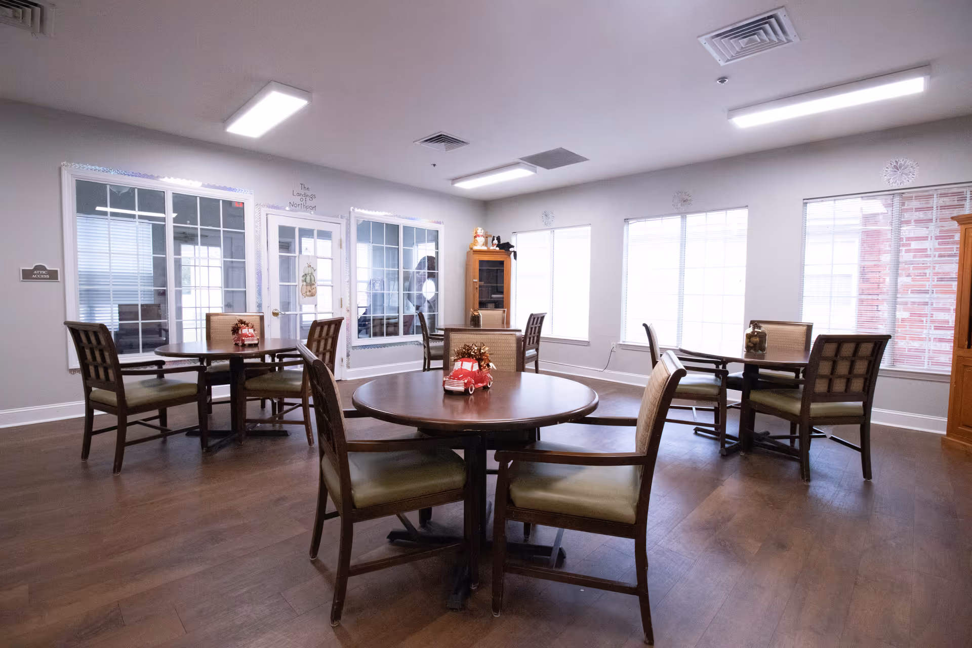 A bright dining room in The Landings of Northport facility with round wooden tables and cushioned chairs arranged neatly. The room has large windows letting in natural light, light-colored walls, and wooden flooring. There are small decorative items on the tables and a wooden cabinet in the corner.