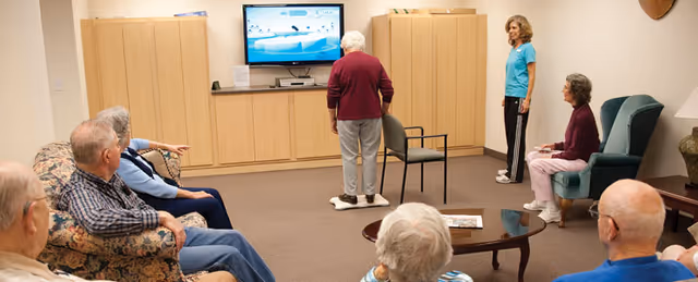 A group of elderly people in a retirement community common room. One elderly woman stands on a balance board facing a television screen, while others sit on chairs and sofas watching her. A staff member in a blue shirt stands nearby, observing the activity. The room has beige walls, carpeted floor, and wooden cabinets along the back wall.