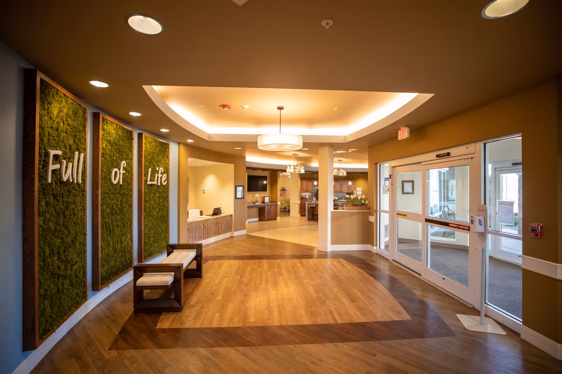 A spacious and well-lit senior living facility lobby area with wooden flooring and a modern ceiling light fixture. On the left wall, there are three vertical green moss panels with the words 'Full of Life' displayed across them. There are two wooden benches with white cushions beneath the panels. To the right, there are glass automatic doors leading outside, and a hand sanitizer dispenser is mounted on a stand near the entrance. The reception desk and a common area with kitchen cabinets and seating are visible further inside.
