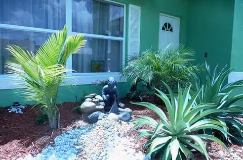 Green facility exterior with front landscaping, palms, rocks, and a garden statue.