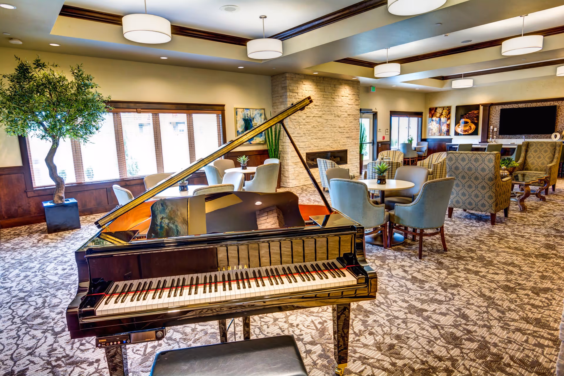 A spacious and well-lit common area featuring a black grand piano in the foreground, several round tables with upholstered chairs, patterned armchairs, a stone fireplace, large windows with wooden trim, and decorative plants. The ceiling has recessed lighting and modern circular light fixtures.