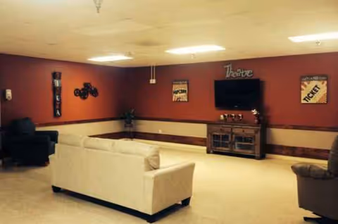 Spacious common room with white couches, a wall TV, and decorative cabinet against maroon walls.