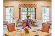 Bright common dining area with round tables set with cups and floral centerpieces and armchairs arranged near large windows.