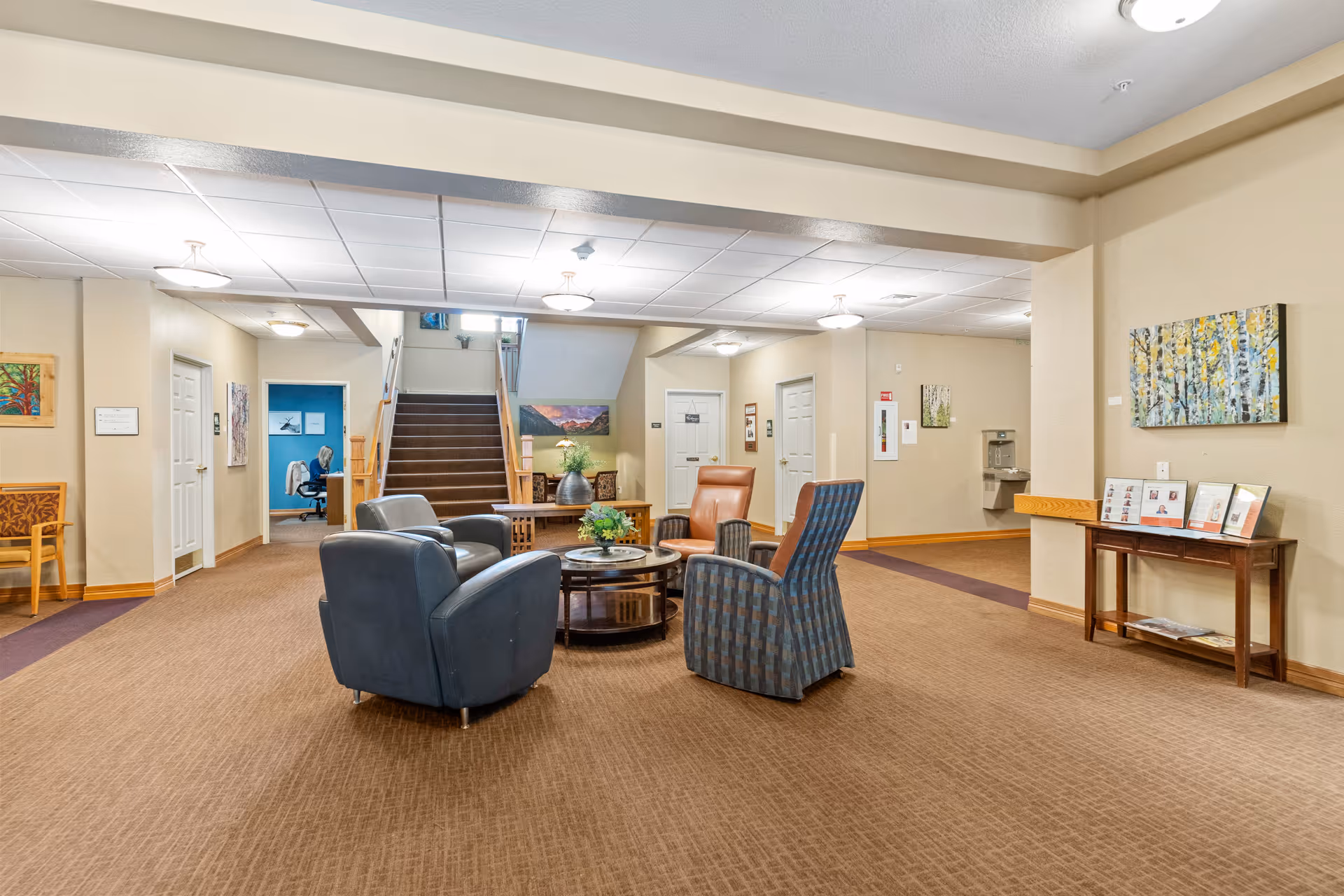 A spacious common area in an assisted living facility with a seating arrangement of four armchairs around a circular coffee table. The room has beige walls, carpeted floors, and ceiling lights. There are stairs in the background leading to an upper floor, a water fountain on the right wall, and various framed artworks and informational displays on the walls.