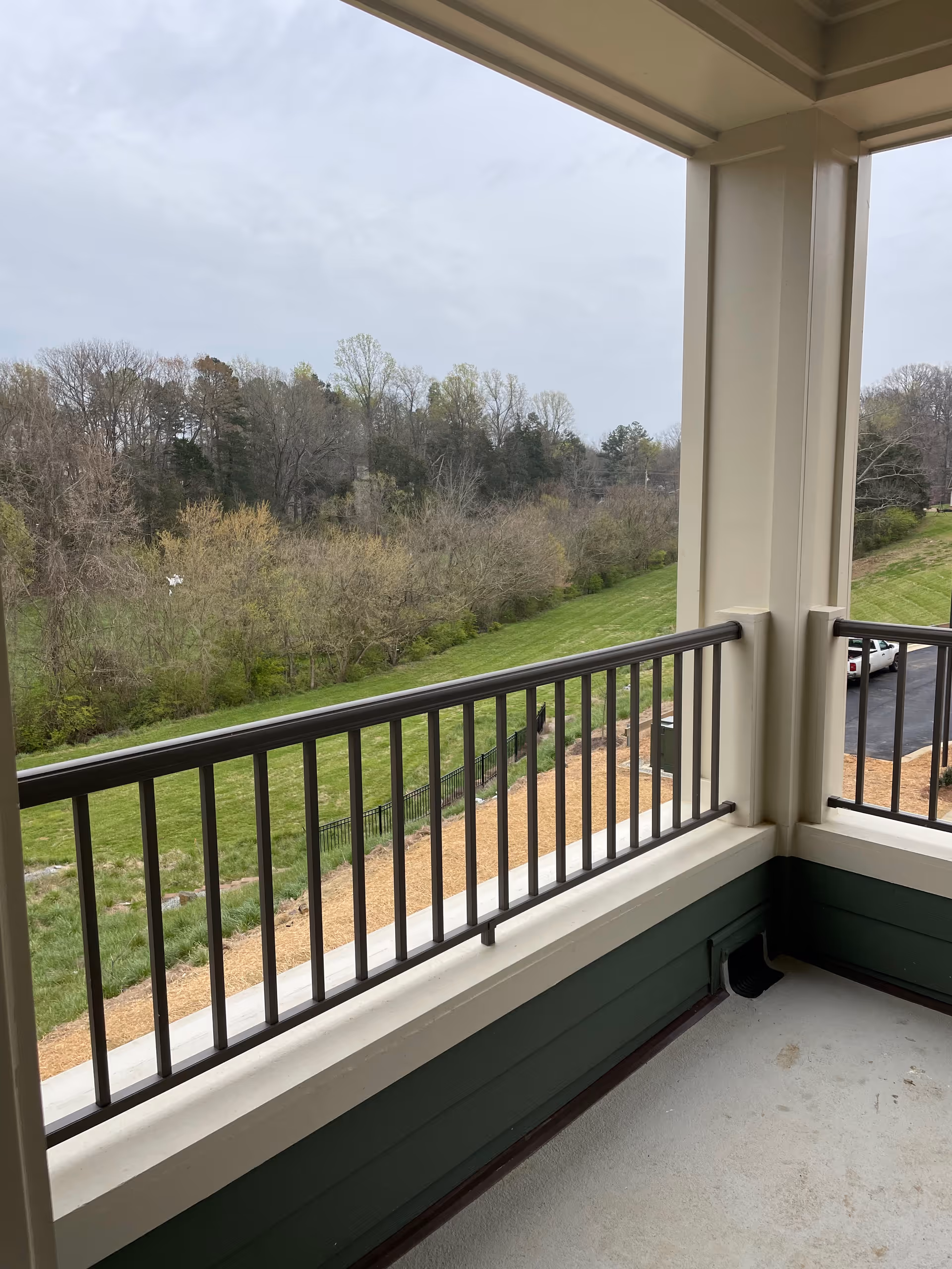 View from a covered balcony with metal railings overlooking a grassy area, trees, and a parking lot with a white vehicle visible in the distance under a cloudy sky.